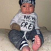 Angel is registered to the contest to win money with this photo: baby, bare_feet, beanie, bed, blanket, child, clothing, facial_expression, hat, indoors, infant, onesie, pants, photography, portrait, sitting, smiling, text_on_clothing, toes, wall