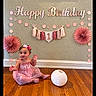 baby, balloon, birthday, birthday_decorations, bunting, celebration, child, happy_birthday, indoors, lantern, paper_flowers, party, pink_dress, portrait, sitting, smiling, toddler, wall, waving, wooden_floor