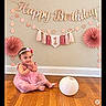 Tessa is registered to the contest to win money with this photo: baby, child, birthday, birthday_decoration, pink_dress, dress, headband, flower, smiling, sitting, indoor, wooden_floor, wall, paper_fan, garland, tassel, lantern, party, portrait, celebration