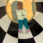 Aaliyah joined the competition — help win amazing prizes! black_and_white, braided_hair, checkered_pattern, child, circle, colorful, cute, frame, fun, hands_on_face, happy, indoor, pants, playful, portrait, seated, smiling, socks, sweatshirt, young