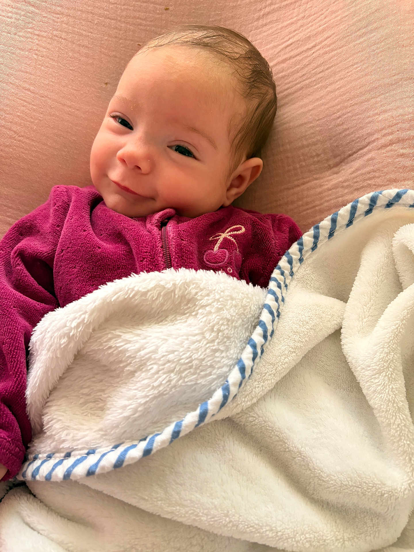 Luana participe au concours pour gagner de l'argent avec cette photo : baby, blanket, purple_clothing, smile, face, infant, soft_texture, cozy, lying_down, cute, white_blanket, blue_trim, pink_background, child, portrait, young, warm_clothing, indoor, peaceful, happy