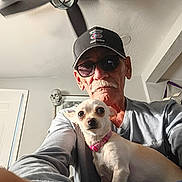 Nena is registered to the contest to win money with this photo: elderly_man, dog, chihuahua, indoor, selfie, sunglasses, cap, ceiling_fan, skull_decoration, american_flag, gray_shirt, pet, pet_collar, portrait, facial_hair, mustache, light, shadow, wall, door