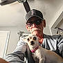 elderly_man, dog, chihuahua, indoor, selfie, sunglasses, cap, ceiling_fan, skull_decoration, american_flag, gray_shirt, pet, pet_collar, portrait, facial_hair, mustache, light, shadow, wall, door