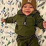 baby, child, smiling, beanie, hat, green_clothing, jacket, pants, blanket, floral_pattern, happy, infant, cute, lying_down, indoor, portrait, face, warm_clothing, cozy, person