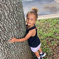 Lyla is registered to the contest to win money with this photo: child, footwear, fun, grass, happy, joy, knee, leisure, people_in_nature, person, plant, sandal, shorts, smile, sneakers, t_shirt, thigh, thumb, toddler, tree