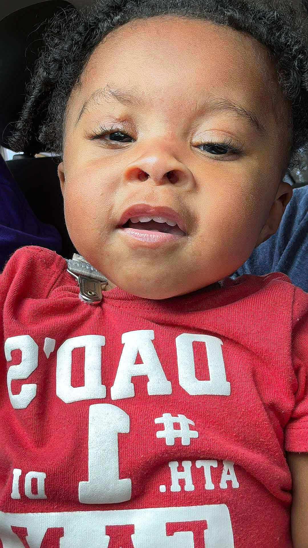 Derrell is registered to the contest to win money with this photo: child, toddler, face, curly_hair, red_shirt, text_on_clothing, close_up, portrait, expression, casual, indoors, cute, young_child, person, fashion, clothing, skin, head, eyes, mouth