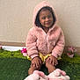 child, toddler, pink_hoodie, yellow_shorts, slippers, bunny_slippers, smiling, sitting, green_grass, artificial_grass, potted_plants, flowers, indoor, cute, happy, portrait, cozy_clothing, wall, hands, feet