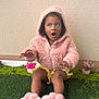 toddler, child, pink_hoodie, fluffy_clothing, bunny_slippers, yellow_skirt, sitting, green_turf, potted_flowers, indoor, expression, curly_hair, cute, gesture, portrait, young_child, playful, cozy, casual, wall_background