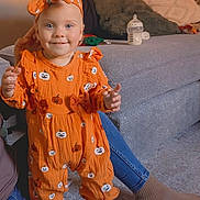 Raven is registered to the contest to win money with this photo: adult_hand, baby_bottle, carpet, child, clothing, couch, cozy, cushions, floor, happy, headband, home, indoor, orange_outfit, person, portrait, pumpkin_pattern, smiling, socks, toddler
