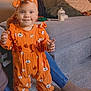adult_hand, baby_bottle, carpet, child, clothing, couch, cozy, cushions, floor, happy, headband, home, indoor, orange_outfit, person, portrait, pumpkin_pattern, smiling, socks, toddler