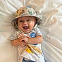 baby, infant, smiling, hat, bucket_hat, tshirt, animal_print, blue_pants, lying_down, happy, child, cute, indoors, white_background, clapping_hands, playful, young, casual_clothing, portrait, joyful