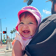 Amelia is registered to the contest to win money with this photo: baby, child, pink_hat, smiling, tattoo, adult, black_shirt, outdoor, sunny, daylight, traffic_light, street, crosswalk, blue_sky, happy, person, holding, arm, face, summer