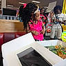 child, birthday, cake, party, pink_outfit, curly_hair, hair_bow, tiara, arcade, restaurant, indoor, people, smiling, celebration, gift_box, birthday_candle, friend, seated, colorful, photograph