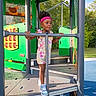 child, toddler, girl, playground, outdoor, smiling, standing, play_structure, safety_rail, pink_visors, floral_outfit, sneakers, blue_shoes, curly_hair, sunlight, trees, park, colorful, recreation, portrait