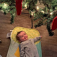 John joined the competition — help win amazing prizes! baby, infant, christmas_tree, christmas_lights, red_stocking, floor, pajamas, mat, holiday, smile, indoors, wood_floor, festive, child, decoration, greenery, cozy, cute, lying_down, celebration