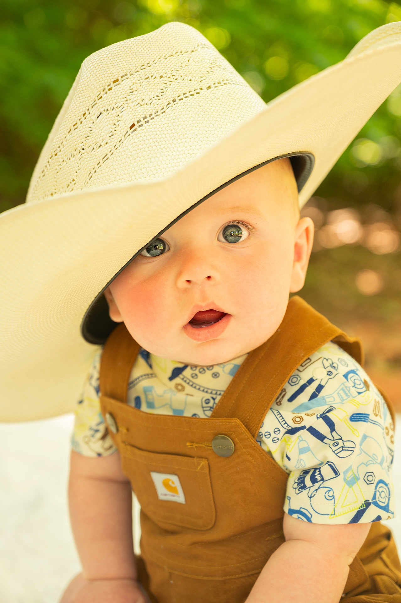 Graesyn is registered to the contest to win money with this photo: baby, child, hat, straw_hat, overalls, patterned_shirt, outdoor, portrait, cute, wide_eyes, surprised_expression, infant, close_up, summer, sunlight, greenery, soft_focus, fashion, casual, adorable