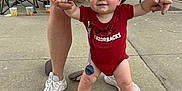 Graesyn is registered to the contest to win money with this photo: adult_support, arkansas_razorbacks, baby, background, beverage_cups, blue_eyes, chair, clothing, concrete, holding_hands, legs, outdoor, parking_lot, people, red_onesie, sidewalk, smiling, sneakers, toddler, walking