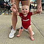 Graesyn is registered to the contest to win money with this photo: baby, toddler, walking, holding_hands, red_onesie, arkansas_razorbacks, adult_support, outdoor, sidewalk, concrete, sneakers, blue_eyes, smiling, people, background, chair, beverage_cups, clothing, legs, parking_lot
