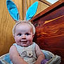 baby, bunny_ears, smile, basket, wooden_background, blue_ears, cute, infant, child, happy, indoors, toy, plush, portrait, sitting, face, closeup, chubby_cheeks, striped_clothing, playful