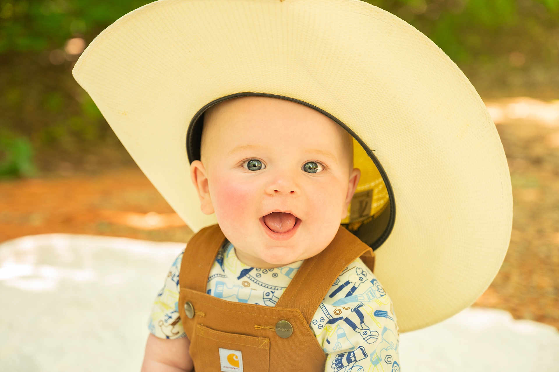 Graesyn joined the competition — help win amazing prizes! baby, background_blur, child, closeup, clothing, cowboy_hat, cute, expression, face, happy, headwear, infant, nature, outdoor, overalls, person, portrait, smiling, sunlight, young