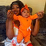 woman, baby, child, orange_clothing, headband, couch, indoor, smile, holding, happy, person, casual_wear, bright_colors, headwear, infant, seated, skin_tone, textile_pattern, home, playful