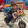 child, motorbike, indoor, amusement_park, ride, toy, smiling, hoodie, pants, colorful, playful, decorations, pink_car, blue_motorbike, lights, fun, playground, young_child, cute, happy