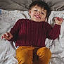 toddler, child, smiling, sweater, maroon, pants, mustard, blanket, lying_down, happy, indoor, face, person, clothing, cute, young, portrait, casual, relaxed, cozy
