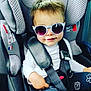 child, toddler, sunglasses, car_seat, seatbelt, smile, white_shirt, black_pants, patterned_pants, hand, hair, face, safety, indoors, cute, baby, fashion, seat, car_interior, accessory