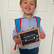 Luccian a rejoint le concours — aidez-le/la à gagner de superbes lots ! announcement, backpack, blue_shirt, boy, casual_clothing, chalkboard, child, door, education, first_day, happy, indoors, jeans, kindergarten, portrait, school, short_hair, sign, smile, standing