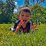 child, boy, grass, flowers, outdoor, nature, trees, blue_sky, clouds, playful, tongue_out, summer, sunlight, meadow, casual_clothing, happy, greenery, person, daytime, fun