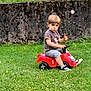 David participe au concours pour gagner de l'argent avec cette photo : boy, casual_clothing, child, daytime, grass, greenery, moss, nature, outdoor, playing, red, serious_expression, shorts, sneakers, steering_wheel, stick, summer, toddler, toy_car, wall