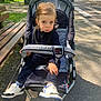 bench, casual, child, clothing, daylight, expression, nature, outdoor, park, pavement, person, seat, shoes, sidewalk, stroller, sunlight, toddler, trees, urban, young_child