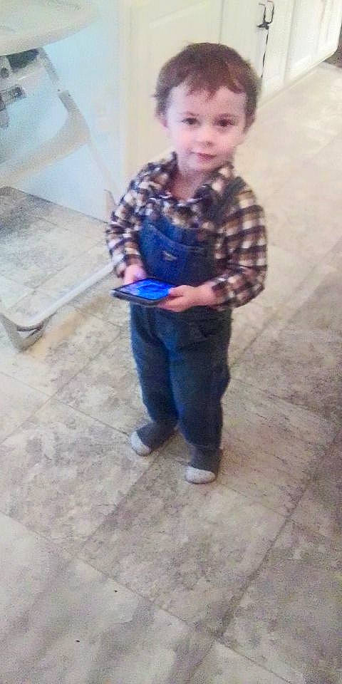 boy, cabinet, casual_clothing, child, curious_expression, denim_overalls, floor_tiles, footwear, high_chair, holding_device, home, indoor, kitchen, person, plaid_shirt, portrait, short_hair, standing, toddler, young