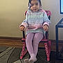 child, girl, braided_hair, beads, sweater, leggings, rocking_chair, pink_chair, indoor, carpet, wood_floor, wall, furniture, portrait, sitting, crossed_legs, calm, casual_clothing, young, home