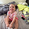 bed, blanket, child, comb, curly_hair, cute, hairbrush, happy, indoors, messy_bed, person, pillow, pink_clothing, playful, room, sitting, smiling, toddler, white_shoes, young_child