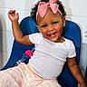 toddler, child, smiling, happy, pink_bow, curly_hair, white_shirt, patterned_pants, blue_chair, toy, sippy_cup, indoor, playful, cute, person, baby, expression, seated, raising_hand, cheerful