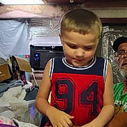 Michael joined the competition — help win amazing prizes! child, boy, toddler, portrait, indoors, sitting, looking_down, short_hair, sleeveless_shirt, jersey, red_clothing, navy_clothing, numbered_shirt, older_person, glasses, hat, blankets, boxes, air_conditioner, home