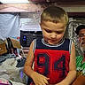 child, boy, toddler, portrait, indoors, sitting, looking_down, short_hair, sleeveless_shirt, jersey, red_clothing, navy_clothing, numbered_shirt, older_person, glasses, hat, blankets, boxes, air_conditioner, home
