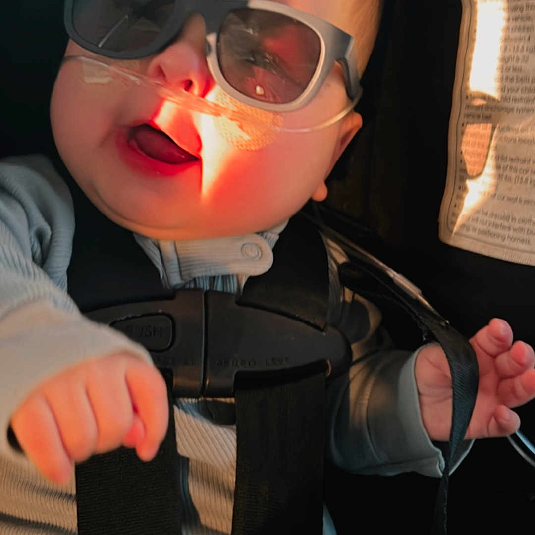 Ander is registered to the contest to win money with this photo: baby, car_seat, child, clothing, cute, expression, face, hand, healthcare, indoor, infant, medical, nasal_cannula, person, safety_harness, seatbelt, smiling, straps, sunglasses, sunlight