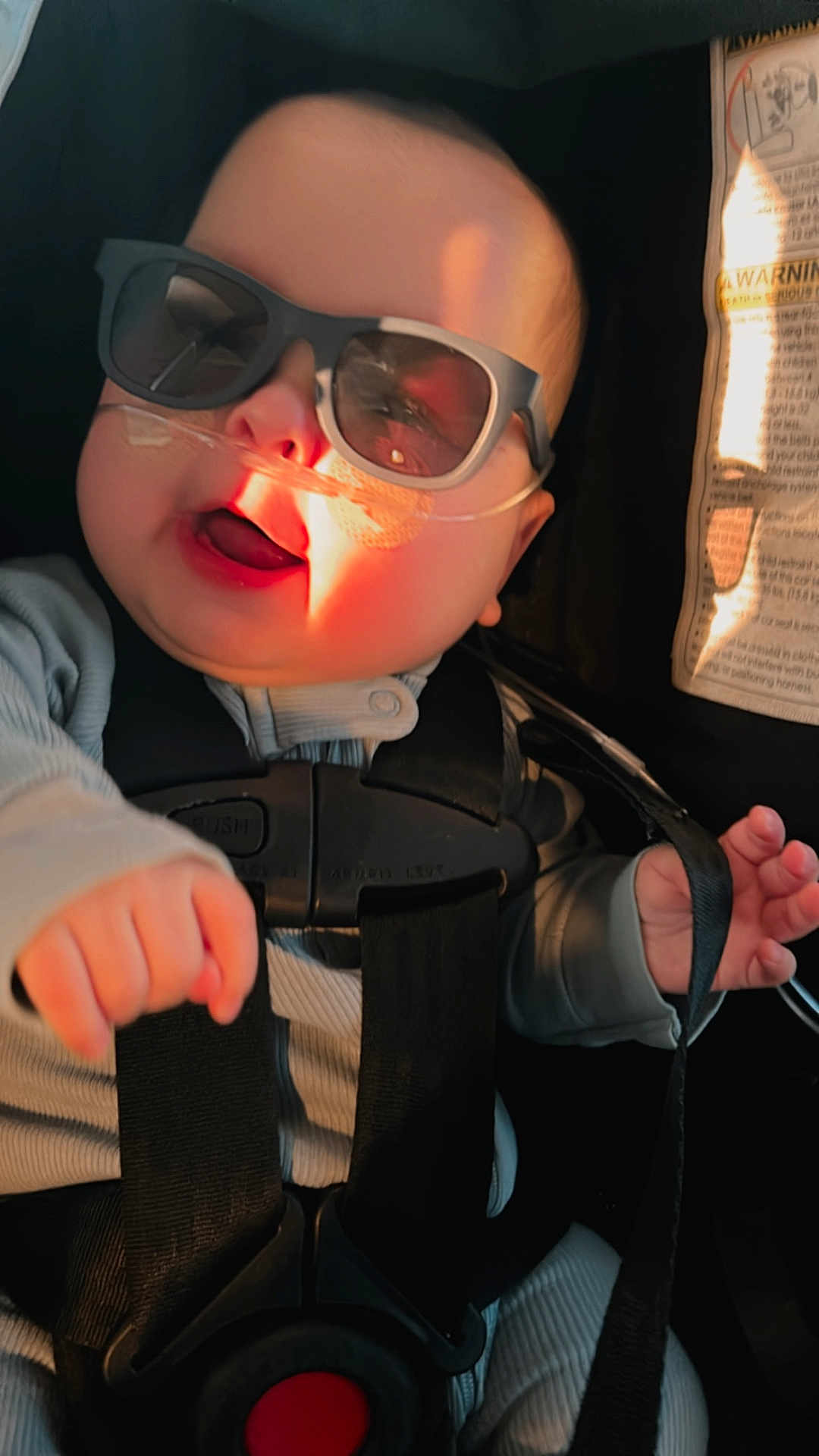 Ander is registered to the contest to win money with this photo: baby, sunglasses, car_seat, nasal_cannula, child, infant, sunlight, smiling, clothing, straps, safety_harness, face, hand, person, indoor, cute, healthcare, medical, seatbelt, expression