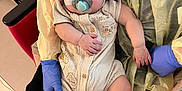 Ander is registered to the contest to win money with this photo: baby, pacifier, sleeping, medical_tape, gloves, handwritten_note, beige_outfit, honeybee_print, socks, infant, child, hospital, chair, yellow_cloth, person, cute, resting, holding, healthcare, comfort