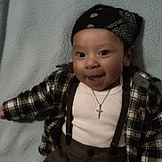 Julian is registered to the contest to win money with this photo: baby, child, smiling, plaid_shirt, bandana, suspenders, cross_necklace, white_shirt, pants, lying_down, blue_blanket, happy, cute, portrait, face, headwear, clothing, infant, person, indoors