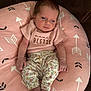 baby, infant, pink_cushion, arrows_pattern, floral_pants, pink_shirt, text_on_clothing, cute, small_feet, tiny_hands, relaxed, indoor, couch, soft_texture, portrait, newborn, child, person, seated, cozy