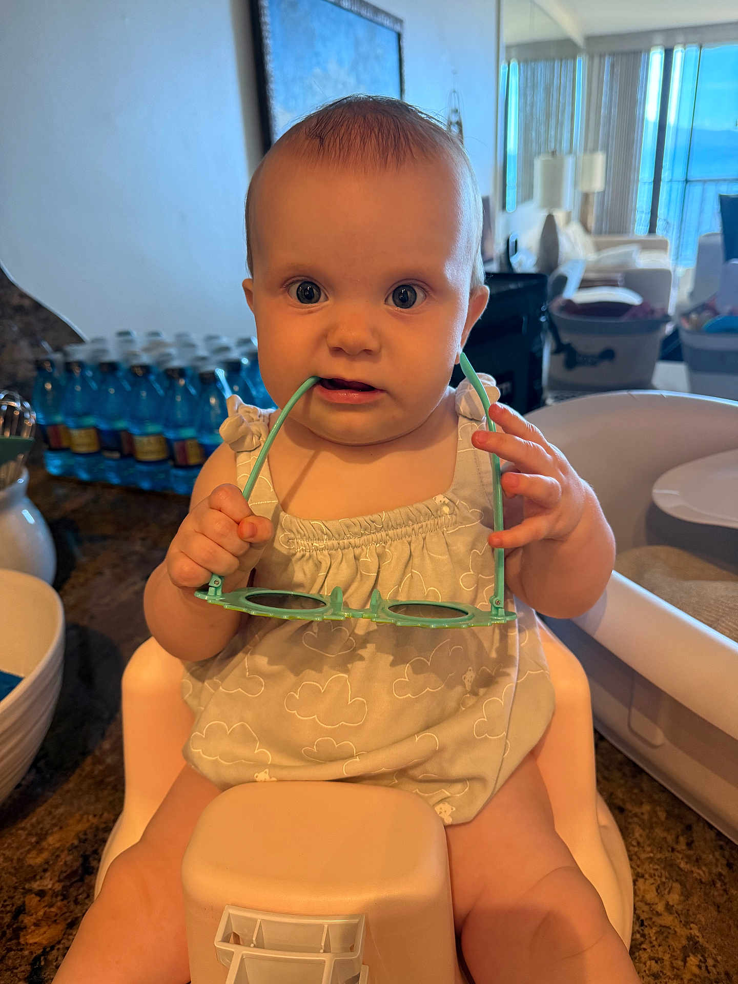 Sloane joined the competition — help win amazing prizes! baby, child, high_chair, sunglasses, kitchen, indoor, person, clothing, furniture, table, bottle, face, hand, curious, seated, infant, expression, light, room, playful