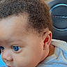baby, child, blue_eyes, curly_hair, car_interior, steering_wheel, camaro, shirt, sun_design, hand, close_up, portrait, infant, seat, young_child, cute, person, indoors, car_seat, expression