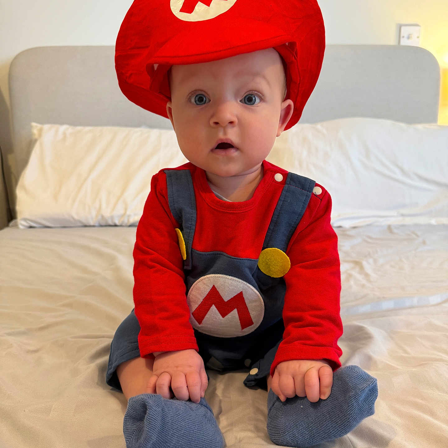 Tommy is registered to the contest to win money with this photo: baby, bed, bedroom, blue_socks, child, clothing, costume, cute, expression, headwear, home, indoors, overalls, person, portrait, red_hat, sitting, soft_lighting, wide_eyes, young_child