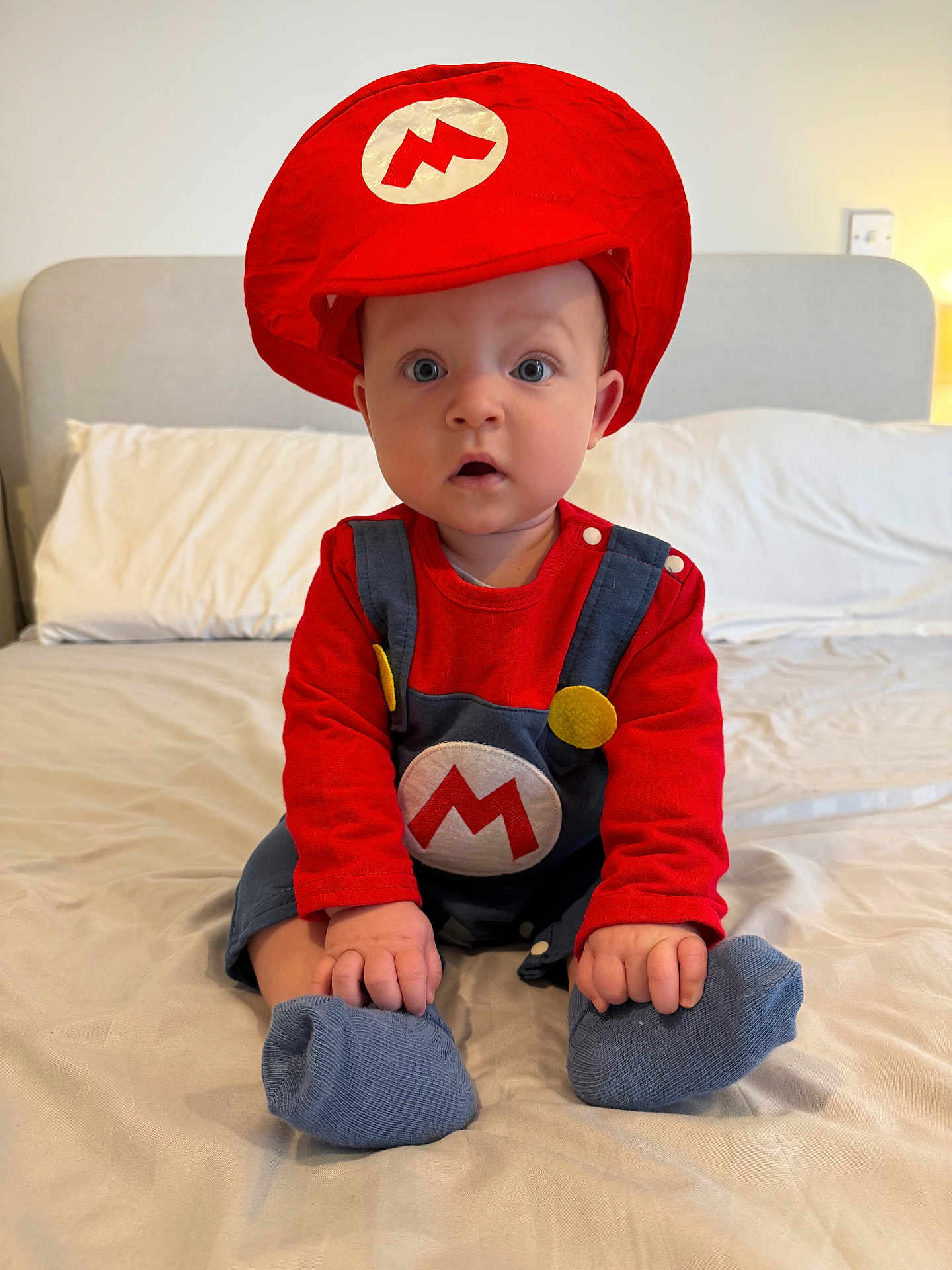 Tommy is registered to the contest to win money with this photo: baby, bed, bedroom, blue_socks, child, clothing, costume, cute, expression, headwear, home, indoors, overalls, person, portrait, red_hat, sitting, soft_lighting, wide_eyes, young_child