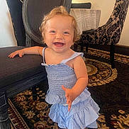 Sophia is registered to the contest to win money with this photo: toddler, child, smiling, dress, indoor, carpet, chair, furniture, happy, person, cute, patterned_chair, floor, young_child, standing, blue_dress, curly_hair, playful, home, interior