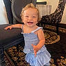 toddler, child, smiling, dress, indoor, carpet, chair, furniture, happy, person, cute, patterned_chair, floor, young_child, standing, blue_dress, curly_hair, playful, home, interior