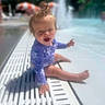 toddler, child, pool, water, swimsuit, sitting, smiling, hair, sunny, outdoor, summer, playful, happy, water_fountain, blue_sky, umbrella, recreation, vacation, fun, baby